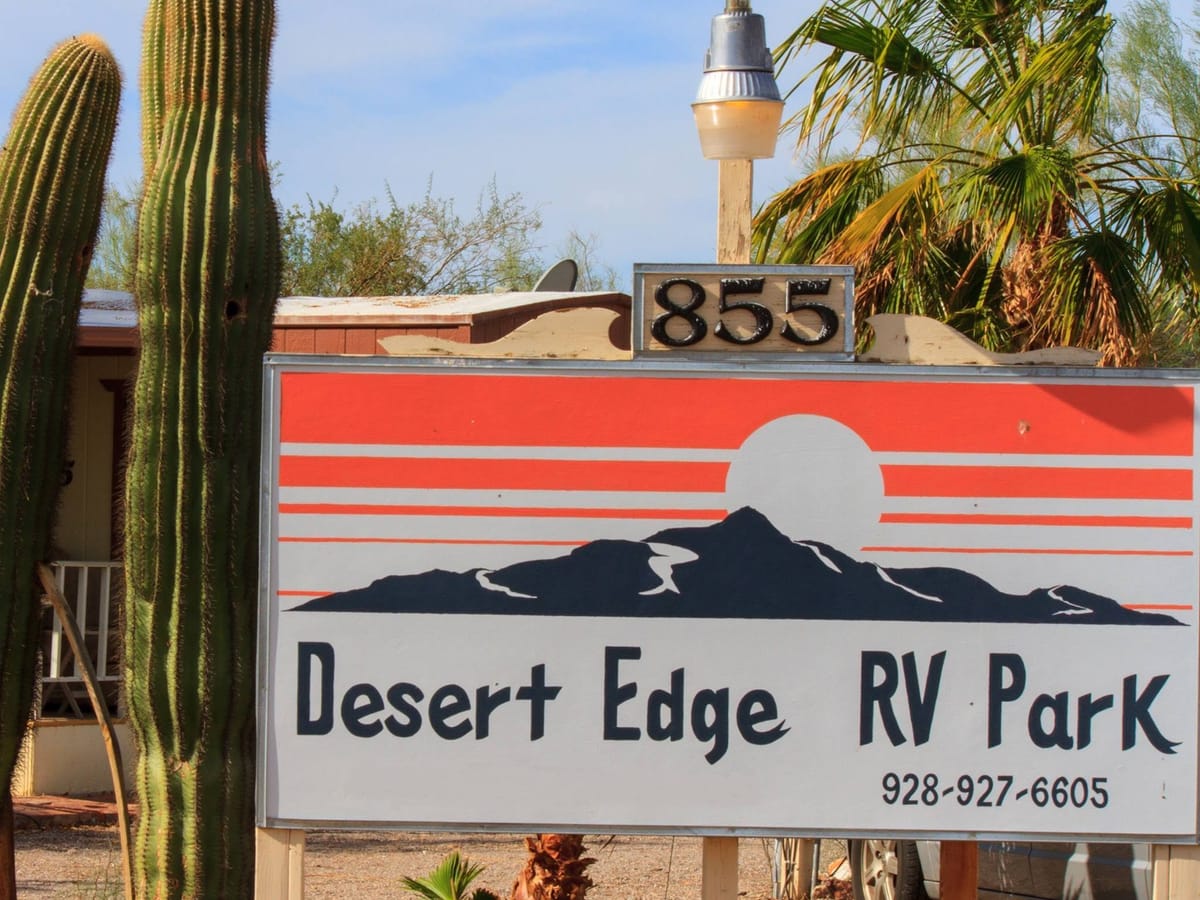 Desert Edge RV Park's entrance sign at 855 E. Rogers Place, painted with a desert sunset, between tall saguaros and a palm